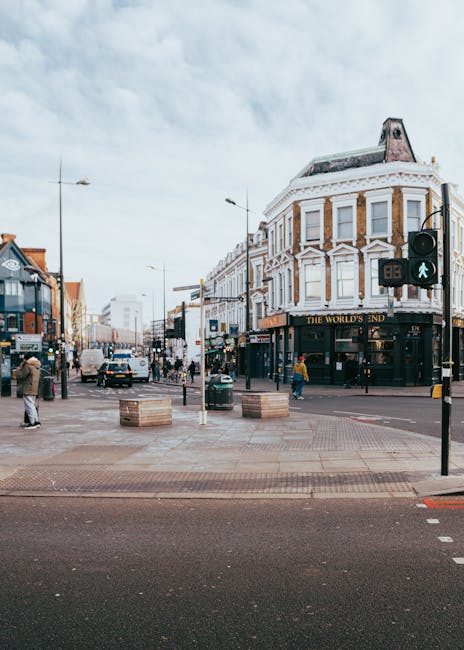 A busy urban street scene in Kentish Town showing a corner building with white decorative brickwork and large windows, housing a pub called 'The World's End' on the ground floor. The street has pedestrians walking on the pavements, some carrying bags or engaged in conversations. A traffic light displays a green pedestrian signal, and vehicles including cars and a double-decker bus are visible on the road. The foreground features a paved pedestrian area with wooden planters and concrete barriers, possibly part of a home relocation project. Overhead, there are streetlights and traffic signals, and the sky is partly cloudy, casting diffuse light over the scene. The setting reflects an active commercial and residential area suitable for house removals and furniture transport, as handled by local removal specialists like Man and a Van Kentish Town.