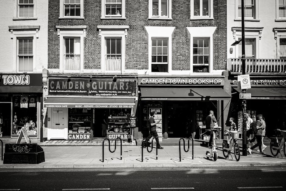 A black and white photograph of a busy street scene in Kentish Town, showing the ground floor façade of a row of commercial buildings with signs for Camden Guitars and Camden Town Bookshop. The storefronts have large glass windows and awnings, one of which displays the shop's name. Several pedestrians walk along the pavement, some carrying bags or backpacks, and appear to be engaged in conversation or browsing. Two bicycles are parked near bike racks in the foreground, while a van is partially visible on the right side, indicating a moving or loading process. The interior of the shops is not visible, but the scene suggests activity related to house removals or furniture transport, consistent with professional moving services provided by Man and a Van Kentish Town. The lighting indicates daytime with natural light illuminating the storefronts and street scene, and the architecture above the shops consists of multiple-story residential buildings with evenly spaced windows and brick facades.