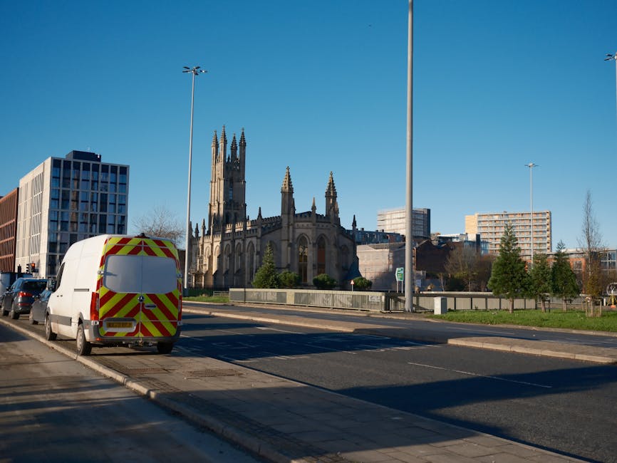 A white van with yellow and red reflective chevron markings parked on a street curb near the entrance to a property, with the back doors closed. The van is positioned alongside several other vehicles parked on the side of the road. In the background, a large historic church with tall, ornate spires and gothic architectural details is visible, surrounded by modern office buildings and trees. The scene is illuminated by natural daylight, with a clear blue sky overhead. This image depicts the loading process in a typical home relocation scenario, highlighting the importance of careful vehicle positioning during a furniture transport or removals service, as provided by Man and a Van Kentish Town.