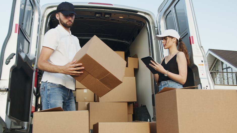 Two individuals are engaged in a home relocation process using a white moving van, parked outside a residential building on a clear day. The man, dressed in a white polo shirt, blue jeans, and a dark cap, is inside the van holding a medium-sized cardboard box, preparing to load or unload it. The woman, wearing a black top, blue jeans, and a white cap, stands beside the entrance of the van, holding a tablet device and checking inventory or planning the packing process. The interior of the van is filled with several packed cardboard boxes of varying sizes, some wrapped in plastic, which are stacked neatly to optimise space during furniture transport. A few larger, flat-pack furniture items are visible among the boxes. The open sliding door of the van reveals a secure loading area with enough room for the transportation of belongings, supporting efficient packing and moving logistics for a home relocation. Surrounding the scene, a paved driveway and a nearby house with a balcony are visible, indicating an organized residential environment connected to professional removals services by Man and a Van Kentish Town, ensuring careful handling of household items during the moving process.