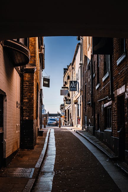 A narrow urban street viewed from underneath an archway with a dark ceiling, showing brick and stone buildings on both sides under clear blue sky. The street surface is a combination of cobblestones and paving slabs, with a slight incline visible. Signs for shops and businesses, including one labeled 'TESCO' and another with 'ARC,' hang from the buildings. Parked cars are seen further down the street, and there are no people or moving vehicles present. The scene is illuminated by daylight, highlighting the textures of the brickwork and street surface. This image could represent the setting for a home relocation or furniture transport process, relevant to house removals services in narrow street environments, as provided by Man and a Van Kentish Town.