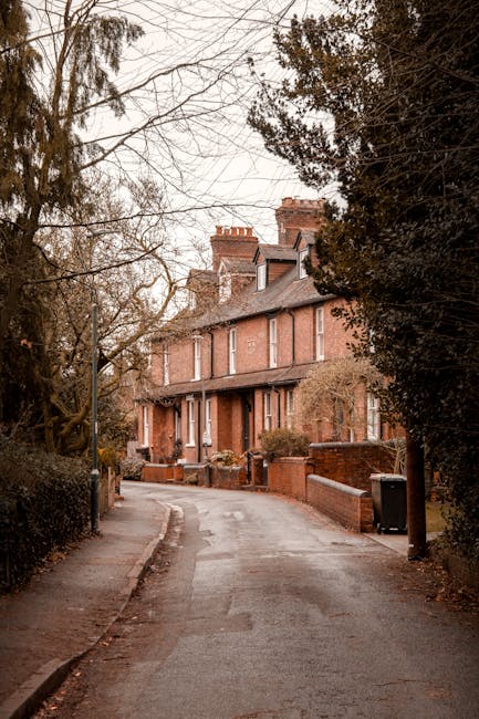 A narrow residential street with a slight incline, lined with mature trees and brick walls on either side. At the end of the street, there is a three-storey red brick house with white window frames and a small front garden. On the pavement near the house, a black trash bin is visible, and the street is empty with no vehicles or people present. The scene is captured during daytime with overcast lighting, highlighting the quiet, typical urban setting suitable for house removals and packing and moving services offered by Man and a Van Kentish Town, who specialize in furniture transport and home relocation on narrow streets such as in Kentish Town.