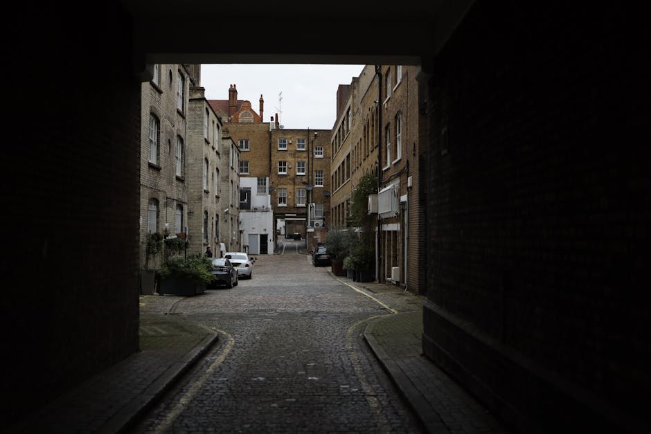 A narrow urban street viewed from underneath an archway with a dark ceiling, showing brick and stone buildings on both sides under clear blue sky. The street surface is a combination of cobblestones and paving slabs, with a slight incline visible. Signs for shops and businesses, including one labeled 'TESCO' and another with 'ARC,' hang from the buildings. Parked cars are seen further down the street, and there are no people or moving vehicles present. The scene is illuminated by daylight, highlighting the textures of the brickwork and street surface. This image could represent the setting for a home relocation or furniture transport process, relevant to house removals services in narrow street environments, as provided by Man and a Van Kentish Town.