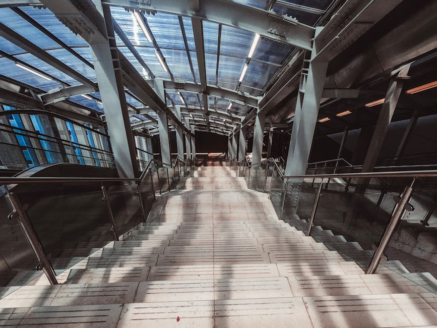 A wide-angle view of a modern, enclosed staircase at Kentish Town West station, illuminated by natural light coming through a glass roof structure. The staircase has light-colored concrete steps with metal railings on both sides and shadows cast by the railing posts. The station's surrounding framework, composed of metal beams and supports, is visible above and alongside the staircase. The environment suggests an urban transit setting, with no visible occupants or moving objects. This indoor station area is part of the preparation for a home relocation or furniture transport process, managed by Man and a Van Kentish Town, and aligns with their removals and moving services, including packing and logistics support for house moves.