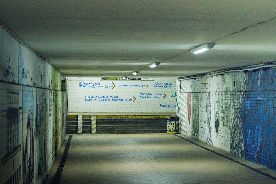 A dimly lit underpass with painted brick walls featuring colorful mural artwork and graffiti. The ceiling has exposed pipes and is illuminated by multiple fluorescent light fixtures. On the far wall, there is a white directional sign with blue handwritten text and arrows indicating local landmarks and distances, such as Zamecky Arad, Jezior Katlov, and The Glassworks Tasice. The floor appears to be tiled, and in the foreground, there are no objects or furniture visible. The scene suggests an urban environment, possibly near a public transport station like Kentish Town West, which relates to home relocation and moving services provided by Man and a Van Kentish Town. This underpass might serve as part of a route during a furniture transport or loading process in a house removal project.