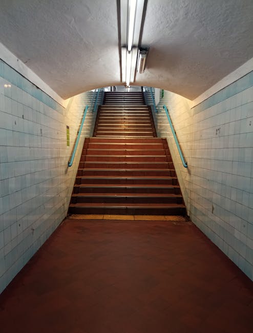 A wide-angle view of a modern, enclosed staircase at Kentish Town West station, illuminated by natural light coming through a glass roof structure. The staircase has light-colored concrete steps with metal railings on both sides and shadows cast by the railing posts. The station's surrounding framework, composed of metal beams and supports, is visible above and alongside the staircase. The environment suggests an urban transit setting, with no visible occupants or moving objects. This indoor station area is part of the preparation for a home relocation or furniture transport process, managed by Man and a Van Kentish Town, and aligns with their removals and moving services, including packing and logistics support for house moves.