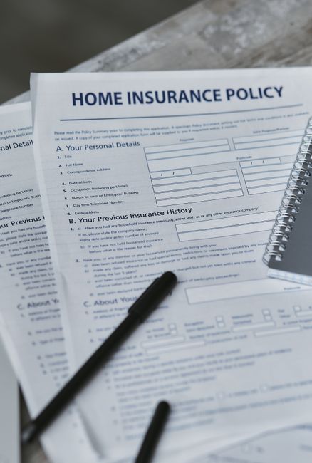 A top-down view of a workspace featuring a clipboard with a printed home insurance policy form, a black pen resting on the clipboard, a clear glass of water, a small gray spiral notebook with a black 