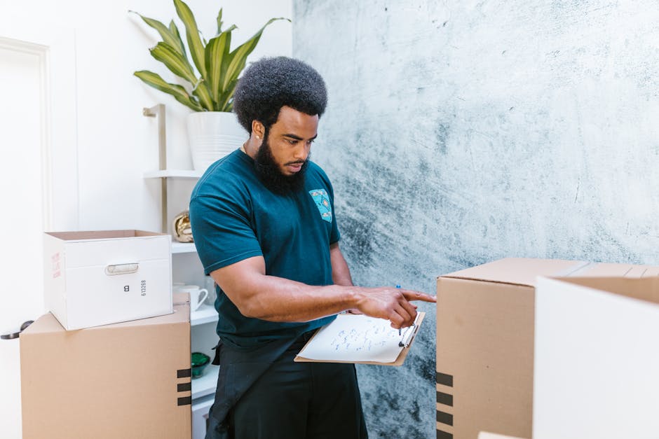 A man with dark curly hair and a beard, wearing a dark teal T-shirt, is inside a room surrounded by packed cardboard boxes. He is holding a clipboard with papers and pointing at the boxes, indicating an inventory or packing checklist process during a home relocation. In the background, there is a white shelving unit with a potted plant on top, and a textured gray wall. The scene depicts the packing and moving stage of house removals, with the man overseeing loading or inventory management in preparation for furniture transport by a professional removals service such as Man and a Van Kentish Town. The boxes are of various sizes, some sealed with packing tape, and positioned on the floor and nearby surfaces, demonstrating an organized packing process in a residential environment.