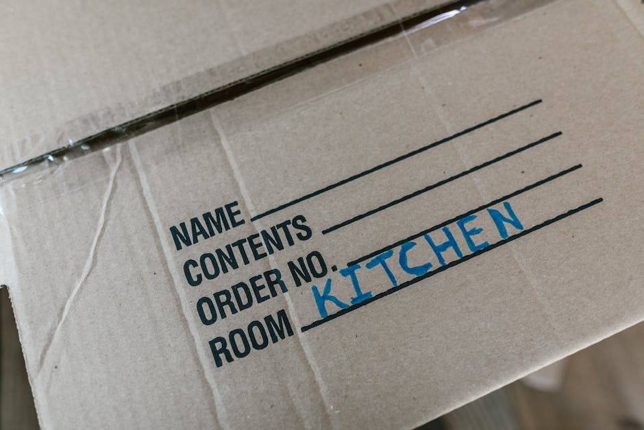 Close-up of a cardboard moving box labeled with black printed sections for 'Name', 'Contents', 'Order No.', and 'Room', with the word 'Kitchen' handwritten in blue marker in the 'Room' field. The box is sealed with clear packing tape and appears to be part of a house removal or relocation process, located inside a property near a doorway, ready for packing or transport. Visible in the image are the cardboard edges and a portion of the top flap, indicating an organized packing process typical of home relocation services provided by Man and a Van Kentish Town, supporting furniture transport and moving logistics.