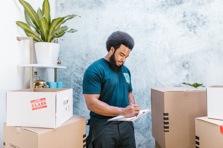 A man with dark curly hair and a beard, wearing a dark teal T-shirt, is inside a room surrounded by packed cardboard boxes. He is holding a clipboard with papers and pointing at the boxes, indicating an inventory or packing checklist process during a home relocation. In the background, there is a white shelving unit with a potted plant on top, and a textured gray wall. The scene depicts the packing and moving stage of house removals, with the man overseeing loading or inventory management in preparation for furniture transport by a professional removals service such as Man and a Van Kentish Town. The boxes are of various sizes, some sealed with packing tape, and positioned on the floor and nearby surfaces, demonstrating an organized packing process in a residential environment.