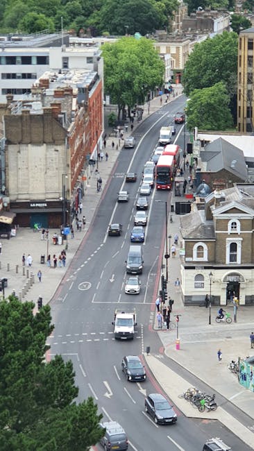 An aerial view of a busy street in Kentish Town featuring multiple lanes of traffic with cars, vans, and buses moving in both directions. On the right side, a row of parked bicycles is seen on the sidewalk near a small commercial building with a rounded roof, adjacent to a larger white structure. The sidewalk is populated with pedestrians and people waiting to cross at the pedestrian crossings. Tall, leafy green trees line both sides of the street, providing shade to the pedestrians. In the background, there are residential and commercial buildings, some with flat roofs and others with traditional brick facades, along with more greenery. The scene captures a typical urban environment suitable for house removals and furniture transport activities by a company such as Man and a Van Kentish Town, illustrating the logistical context of moving services in a bustling city area.