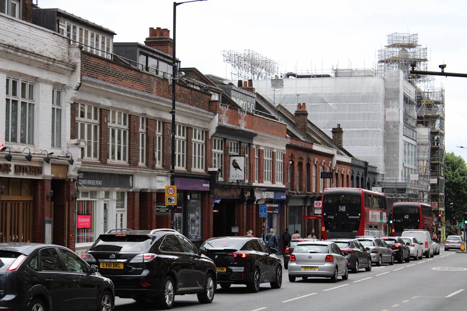 An aerial view of a busy street in Kentish Town featuring multiple lanes of traffic with cars, vans, and buses moving in both directions. On the right side, a row of parked bicycles is seen on the sidewalk near a small commercial building with a rounded roof, adjacent to a larger white structure. The sidewalk is populated with pedestrians and people waiting to cross at the pedestrian crossings. Tall, leafy green trees line both sides of the street, providing shade to the pedestrians. In the background, there are residential and commercial buildings, some with flat roofs and others with traditional brick facades, along with more greenery. The scene captures a typical urban environment suitable for house removals and furniture transport activities by a company such as Man and a Van Kentish Town, illustrating the logistical context of moving services in a bustling city area.
