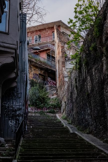 A narrow outdoor staircase made of concrete steps, situated between a graffiti-covered building on the left and a steep, rocky embankment on the right, leading upward to a residential apartment building with a pink exterior and a small balcony visible at the top. The staircase is surrounded by urban landscape elements including a metal pole with signs and wires attached near the top, some greenery growing along the embankment, and a weathered appearance of the surrounding structures. The scene is captured in natural daylight, highlighting the textured surfaces of the stairs and buildings. This setting illustrates a typical access route used during house relocations or moving logistics in challenging urban environments, such as those managed by Man and a Van Kentish Town, specialising in removals with narrow access considerations.