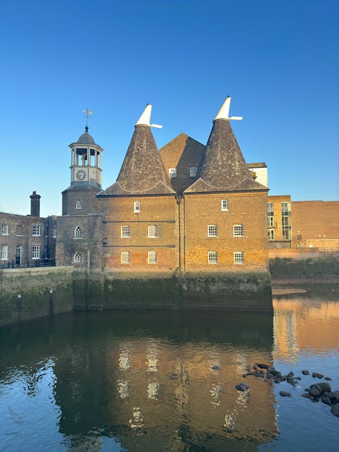 Photograph of a historic brick building with two conical turrets topped with white windmills, situated alongside a river. The building features small, evenly spaced windows and a clock tower with a weather vane on top. The reflection of the building is visible in the calm water, which contains some rocks near the riverbank. The sky is clear and blue, indicating daytime. The scene captures the architecture and setting typical of a heritage site involved in home relocation or building preservation, relevant to house removals and furniture transport services. This image exemplifies the type of location that Man and a Van Kentish Town may work with for deliveries or moving logistics in urban or historic environments.
